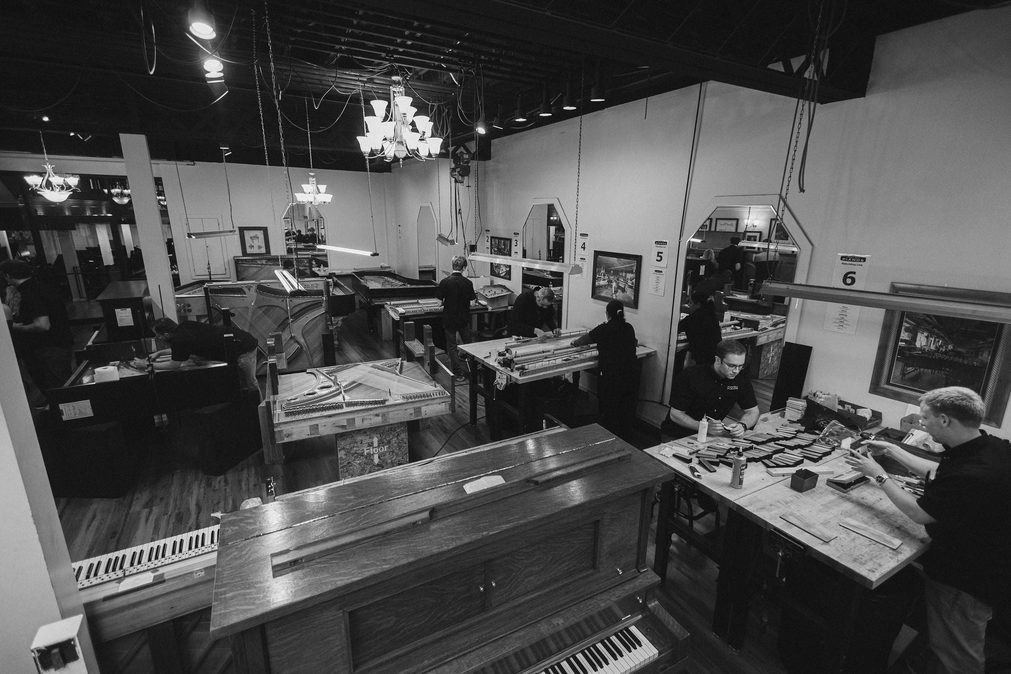 Black and white photo of heirloom upright and grand pianos at various stages of piano restoration (open exposed piano plates, strings, keys), in the interior shop with piano technicians working with equipment at workshop tables in the Brigham Larson Pianos family heirloom piano restoration shop in Orem, UT. 