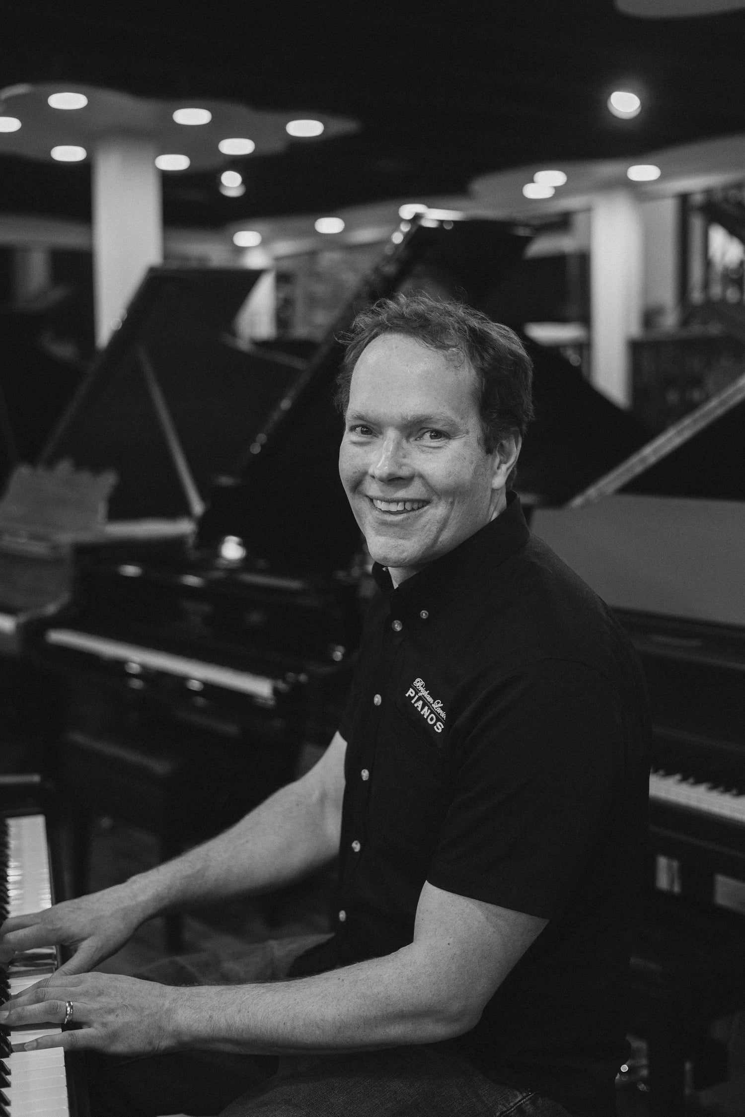Registered Piano Technician Brigham Larson, sitting at a Hailun Grand piano in a showroom setting with several grand pianos in background at Brigham Larson Pianos piano sales showroom in Orem, Utah.