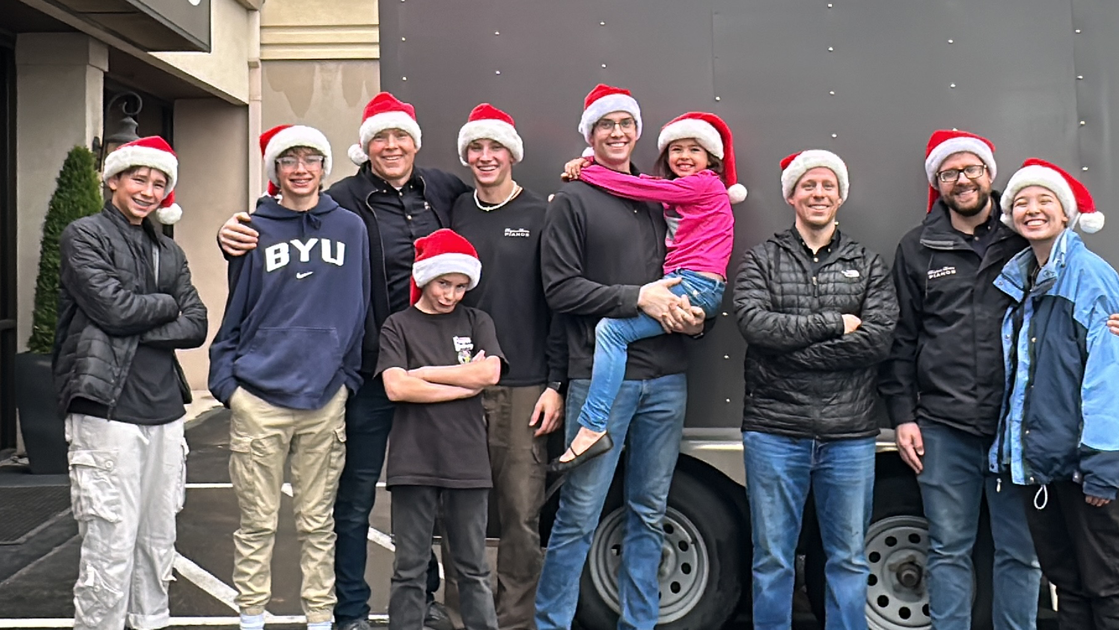 Group of Brigham Larson Pianos piano technicians dressed up for Christmas Eve Piano Deliveries, wearing Christmas hats in front of a piano delivery truck with text overlay.