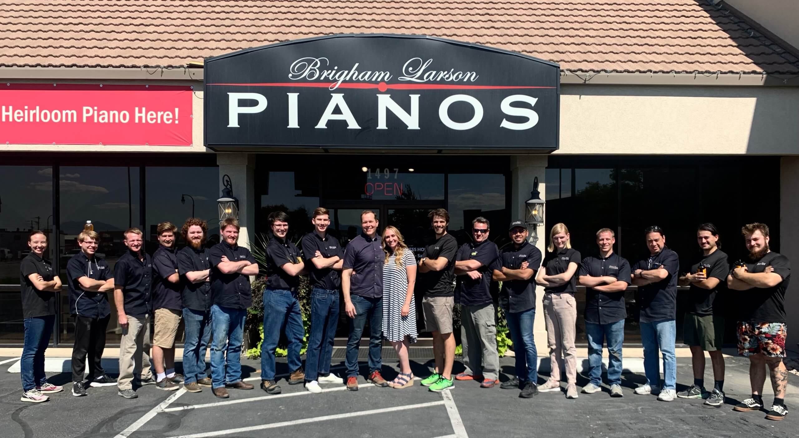 Brigham Larson Pianos team of expert piano rebuilders, tuners, movers, vintage player restorers, refinishers, piano sales, piano keytop restoration, piano marketing and more! The team is standing in a line with their arms crossed, happily smiling, with Brigham and wife Karmel Larson standing in the middle smiling, in front of the Brigham Larson Pianos storefront showroom and restoration shop in Orem, UT, on a sunny day with flower pots and welcoming open sign in the doorway.