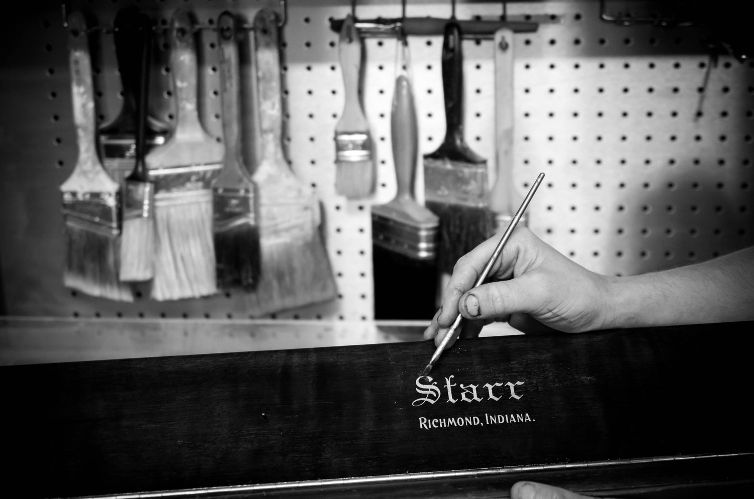 Hand holding a paintbrush near a pegboard with various brushes and a Starr branded piano board being refinished in satin black at Brigham Larson Pianos family heirloom piano restoration shop in Orem, UT.