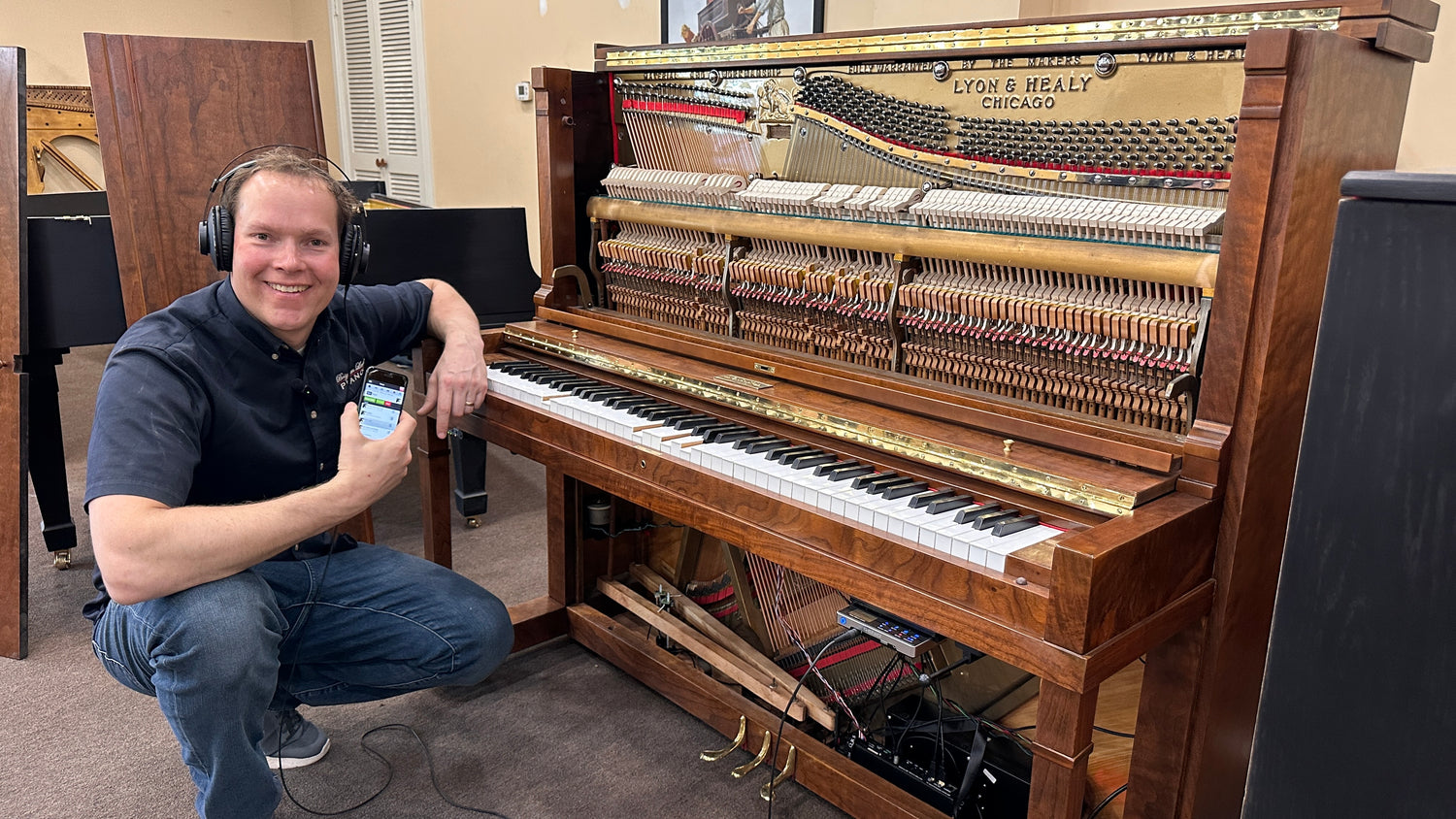 Brigham Larson with headphones and smartphone next to a piano in a room with other pianos, showing the QRS piano player and silent play system installed on a freshly restored heirloom upright Lyon & Healy piano. 
