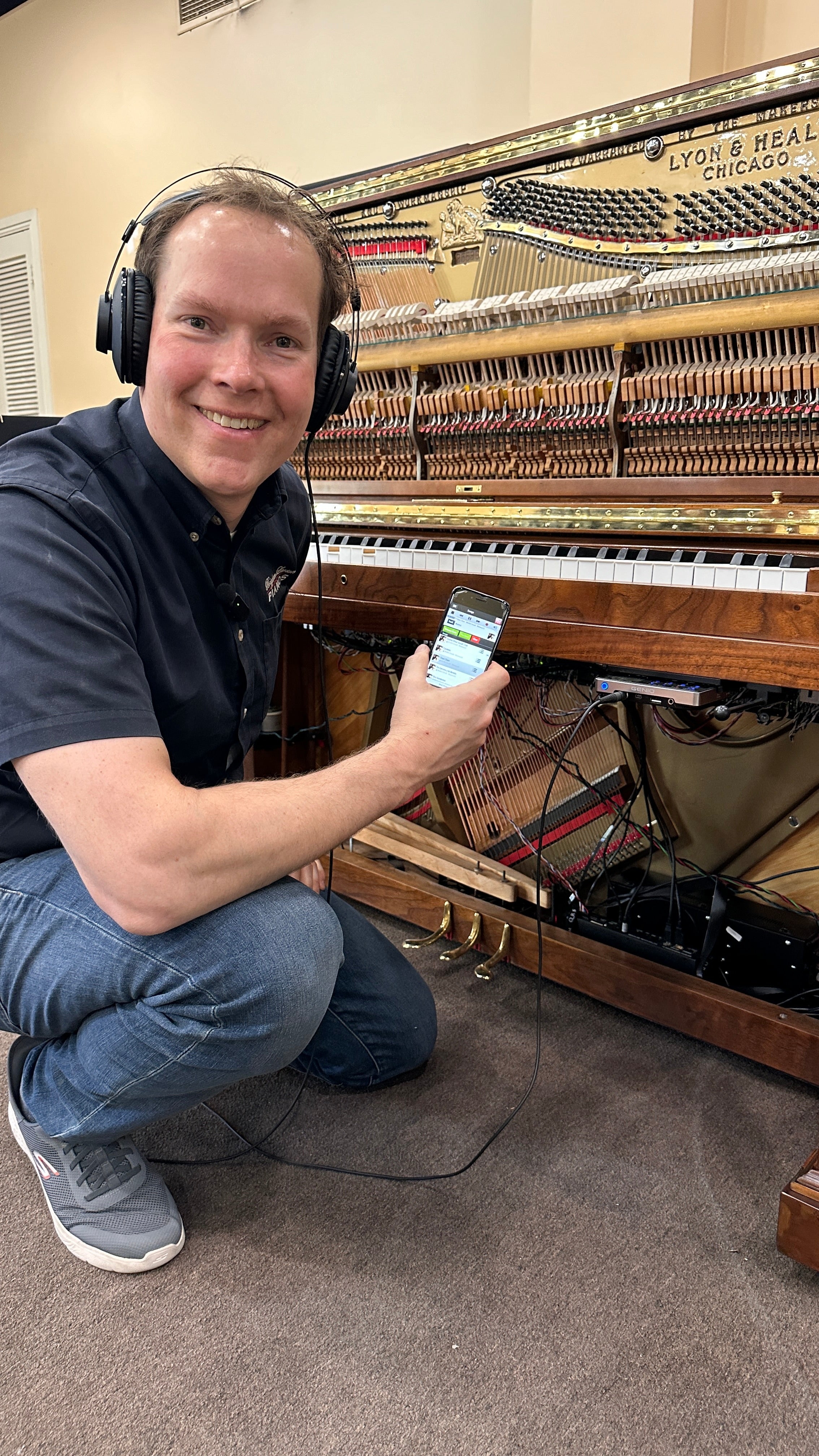Brigham Larson with headphones and smartphone in front of an open upright piano with hammers, strings, and player system QRS showing wires. 