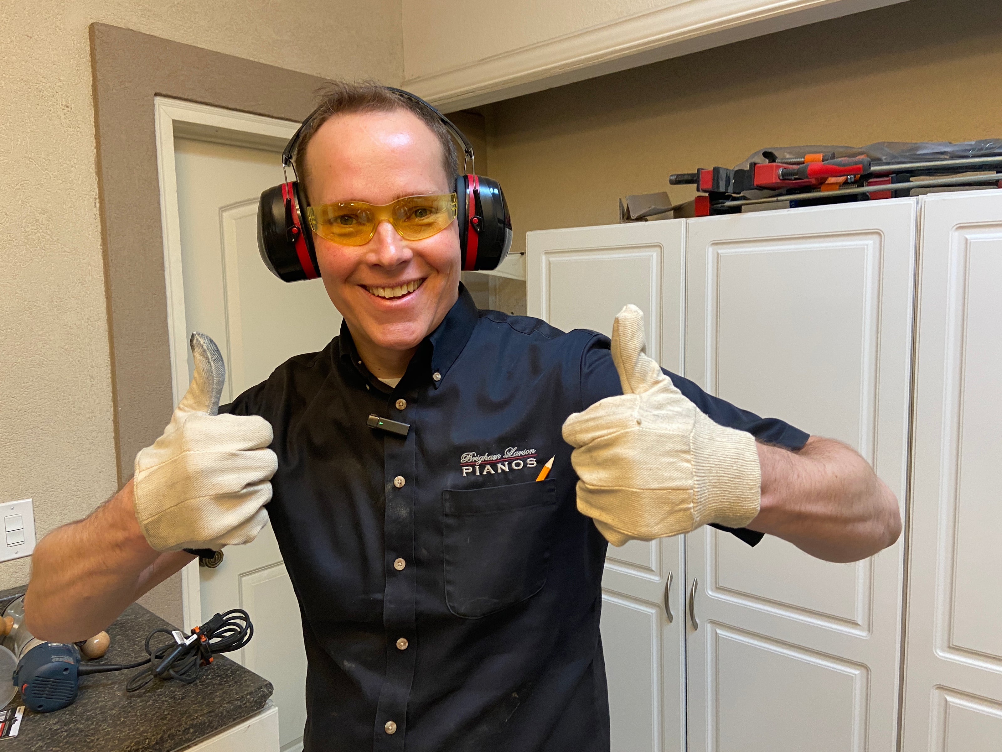 Brigham Larson wearing ear protection and safety glasses, giving a thumbs-up gesture in a piano restoration shop room with tools at Brigham Larson Pianos family heirloom piano restoration shop in Orem, UT.