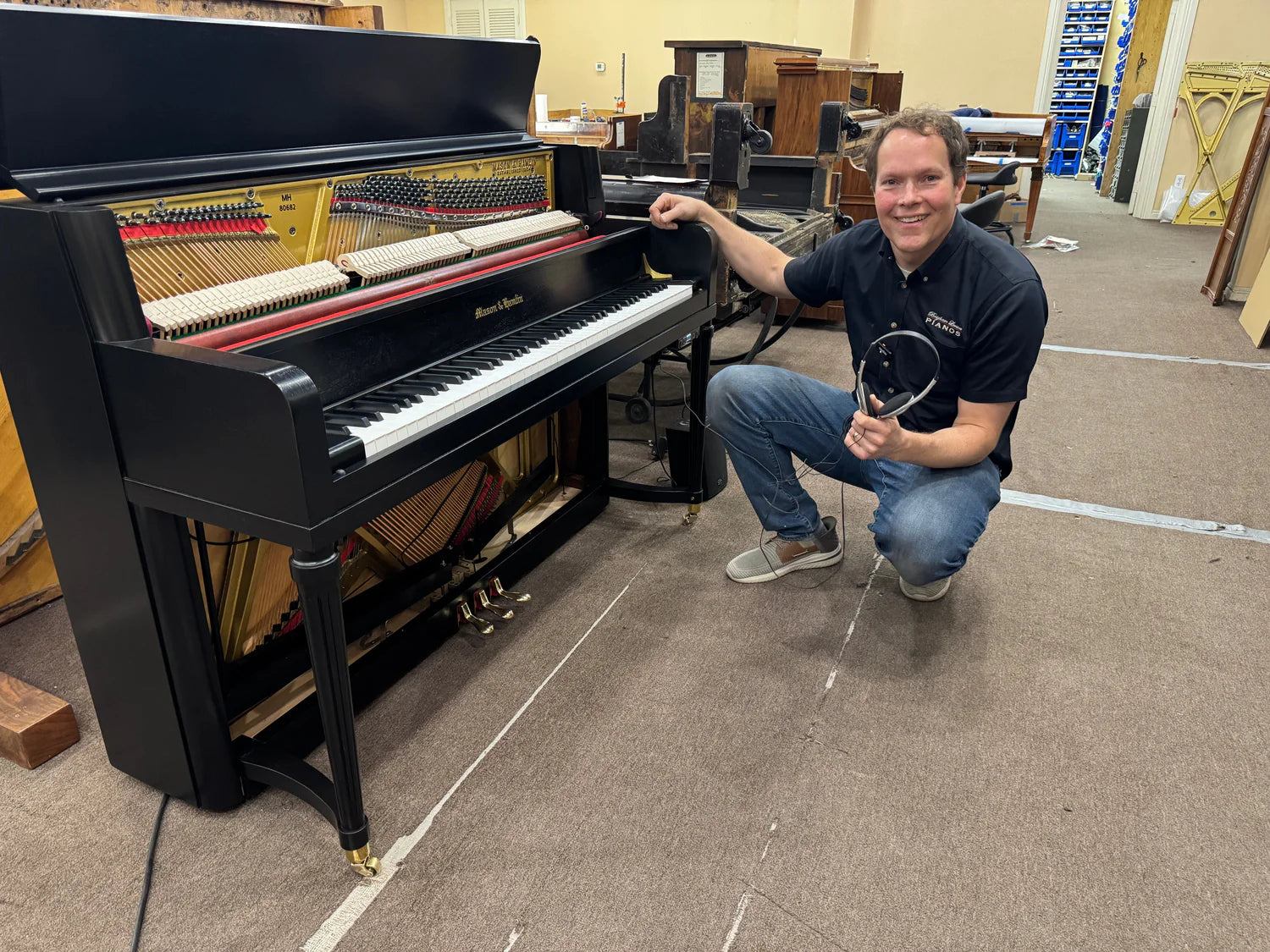 Load video: Brigham holding headphones demonstrating a silenced Rebuilt Heirloom 1977 Mason &amp; Hamlin Console black satin rebuilt and refinished piano.