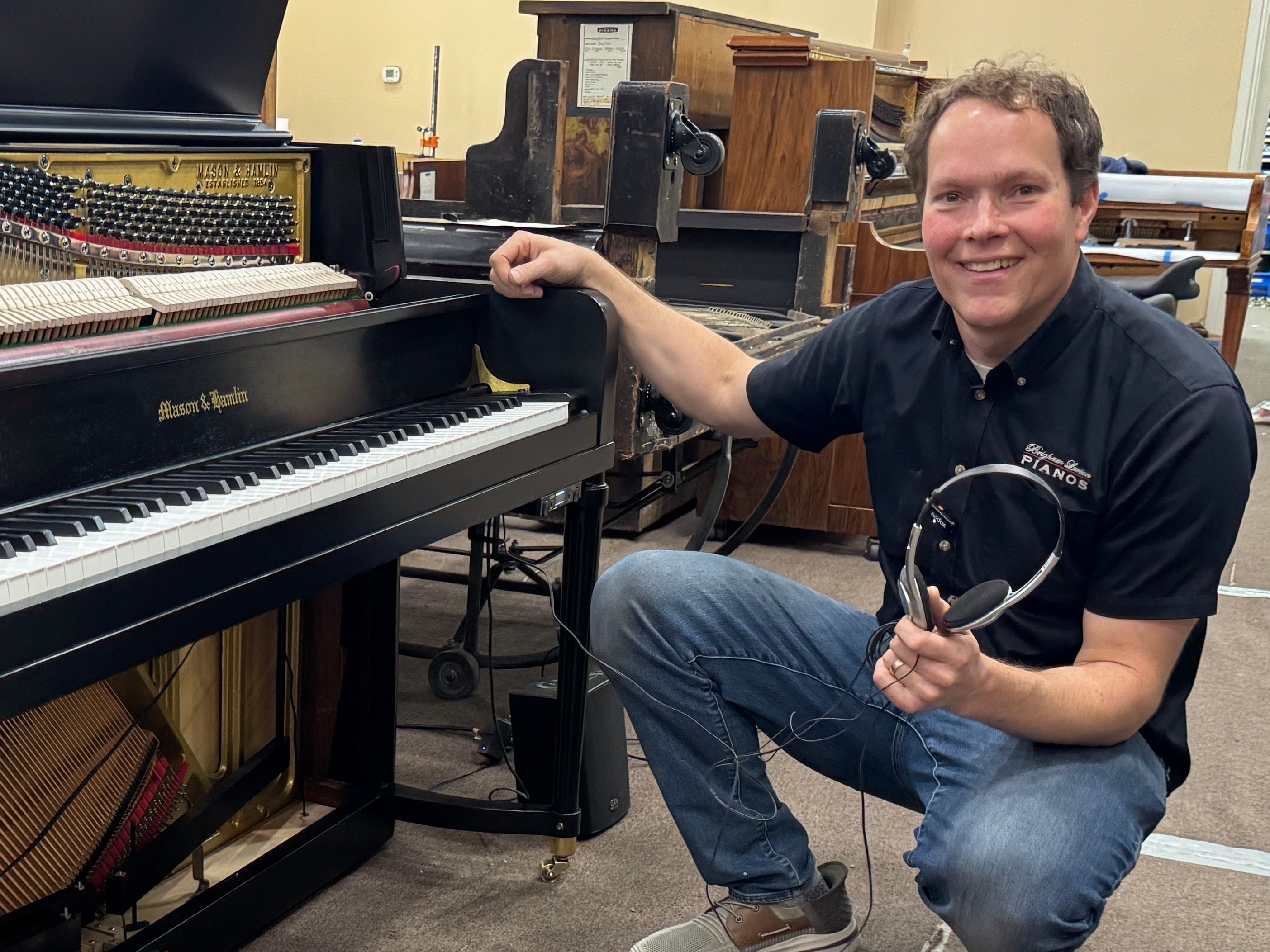 Brigham Larson kneeling next to a piano in a showroom setting holding headphones to a Mason & Hamlin silent play system at Brigham Larson Pianos piano restoration shop.