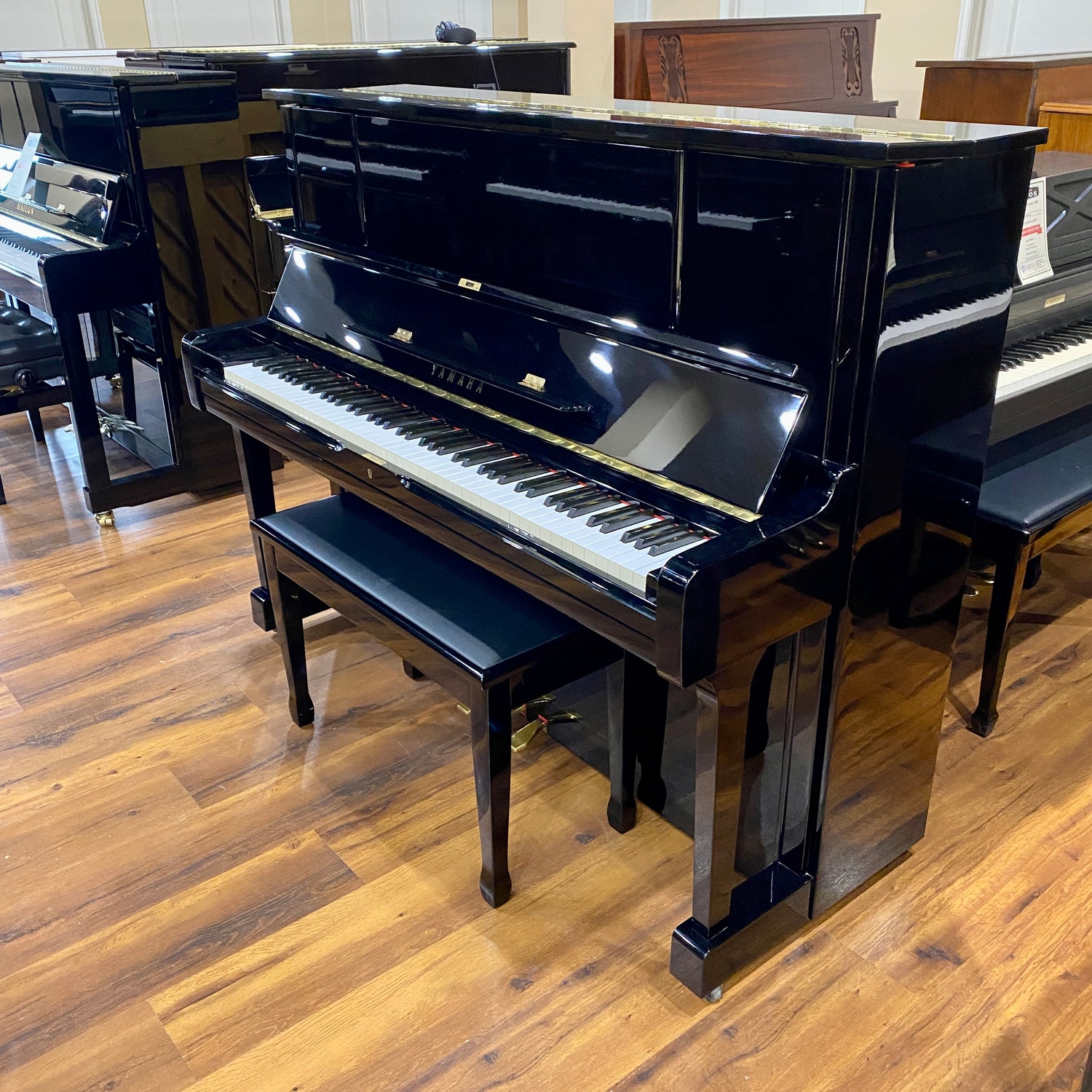 Polished Black Yamaha UX upright piano in a piano sales floor showroom setting with wooden flooring.