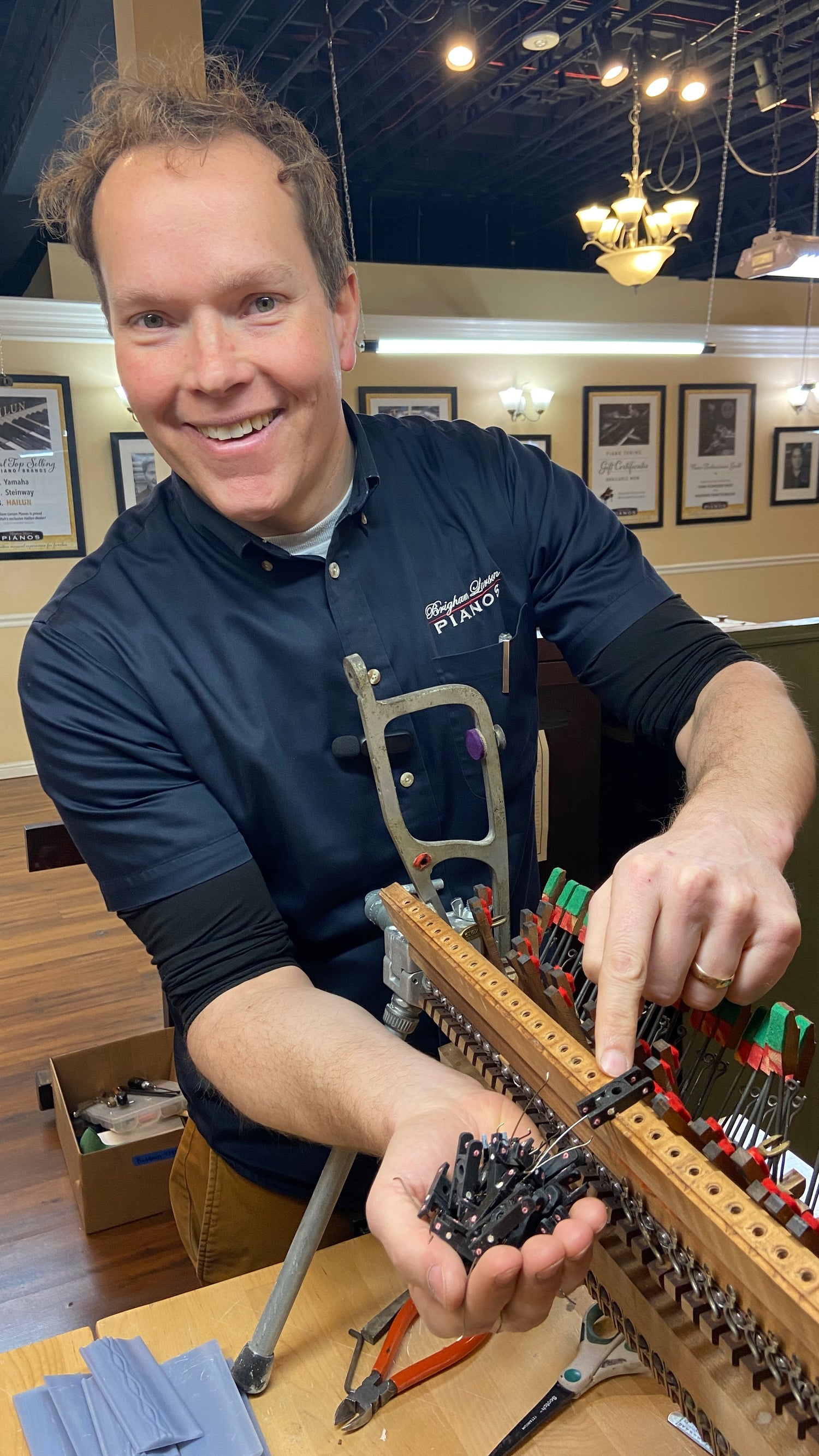 Brigham Larson registered piano technician holding a model train set with tools on a table with 3D printed piano flanges at Brigham Larson Pianos in Orem, Utah piano heirloom piano restoration shop.