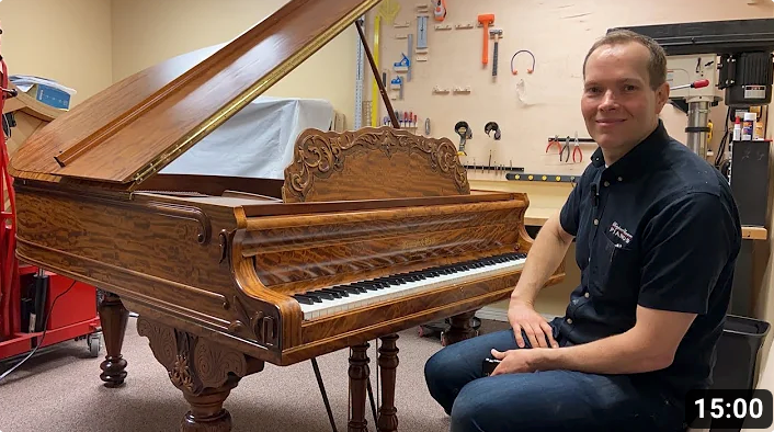 Brigham Larson with restored grand piano ornate cabinetry in piano shop with tools hanging on wall.