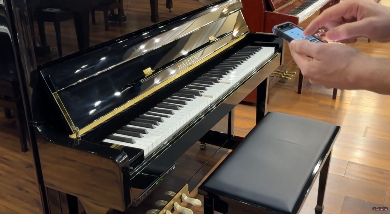 Brigham Larson using a smartphone next to an upright Hailun piano in a piano showroom with wooden flooring at Brigham Larson Pianos in Orem, Utah.
