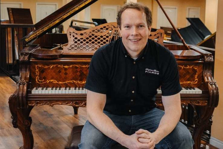 Brigham Larson sitting on a piano bench in front of a fully rebuilt and refinished Steinway grand piano with beautiful woodwork, at Brigham Larson Pianos in Orem, UT.